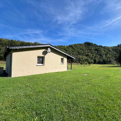 Ein einfaches Haus inmitten einer grünen Wiese. Im Hintergrund sind sanfte Hügel und ein klarer blauer Himmel zu sehen.
