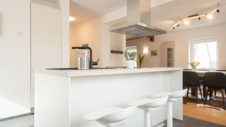 Een moderne keuken met een witte keukenblok en hoge zitplaatsen. De ruimte is licht en vriendelijk ingericht, met een eethoek op de achtergrond.