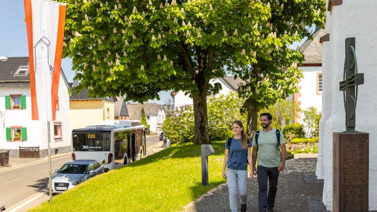 Two hikers are walking along a paved path past a white chapel; an Eifelsteig hiking bus is parked at the side of the road next to it. In the background, green trees, village houses, and a flag frame the sunny village scene.