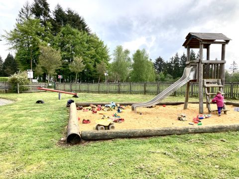 Ein schöner Spielplatz mit einer Rutsche und einer Sandkiste. Im Hintergrund sind Bäume und eine Wiese zu sehen.