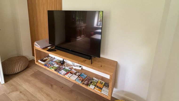 Un téléviseur moderne sur une étagère en bois, remplie de différentes revues. À côté se trouvent des coussins confortables.