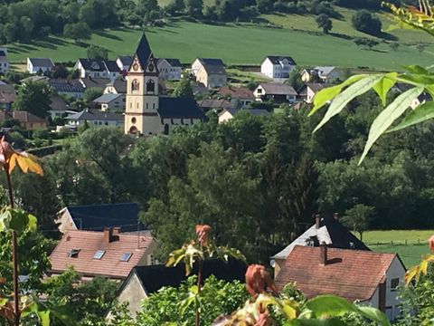 Blick auf ein Dorf mit Kirche und Häusern, umgeben von grünen Hügeln und Bäumen.