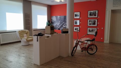 Ein heller Ausstellungsraum mit einem weißen Podest und retro Möbeln. An der Wand hängen verschiedene Fotografien, während ein rotes Wanddesign einen modernen Akzent setzt.