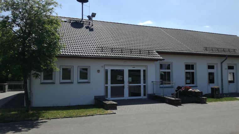 A modern building with a gray roof and numerous windows. In the foreground, there is a paved area and some trees.