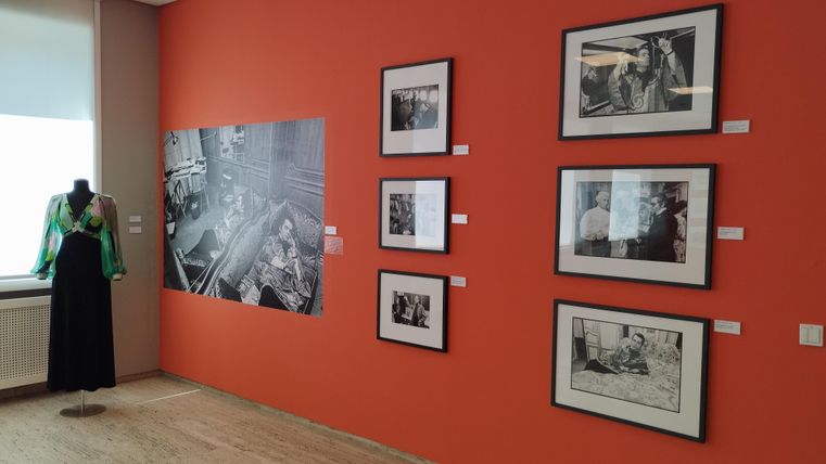 An exhibition room with orange-colored walls. Black-and-white photographs hang on the wall, and on the left, there is a mannequin with a green top.
