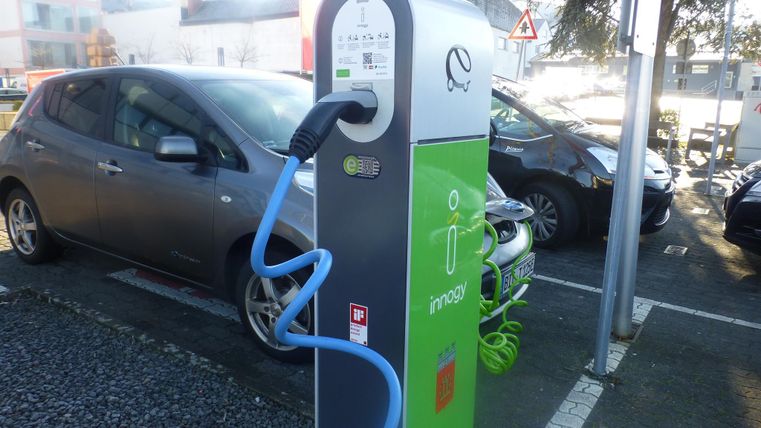 Eine Ladestation für Elektroautos steht auf einem Parkplatz. Neben der Station ist ein graues Auto geparkt.