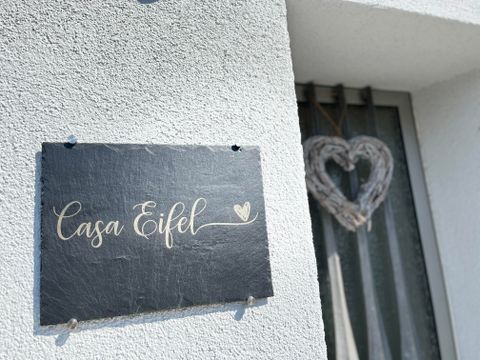 Ein schwarzes Schild mit der Aufschrift "Casa Eifel" hängt an einer weißen Wand. Neben dem Schild hängt ein herzförmiges Deko-Element.