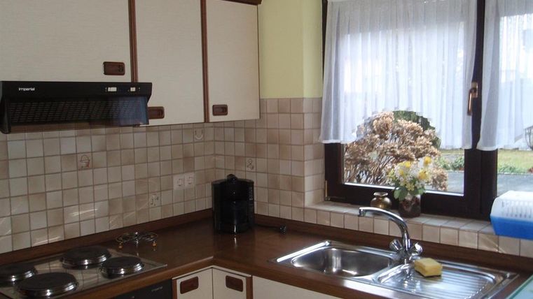 Eine helle Küche mit Holzoberflächen und hellen Fliesen. Am Fenster stehen Blumen und das Licht scheint herein.