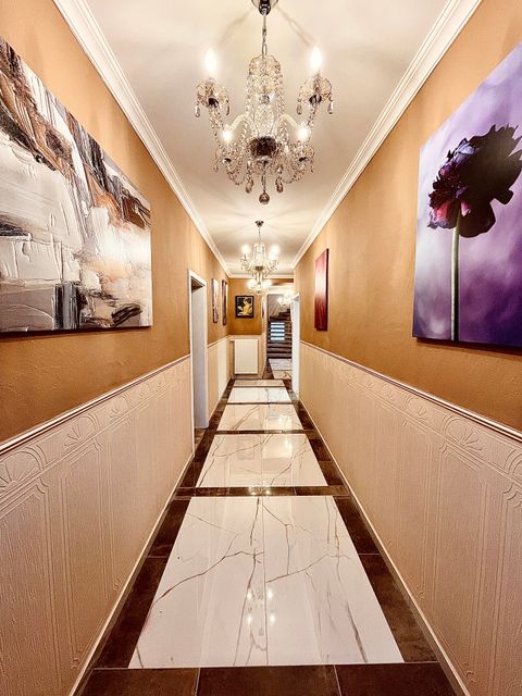 An elegant hallway with a shiny marble floor and tasteful wall paintings. At the end of the hallway hangs a beautiful chandelier.