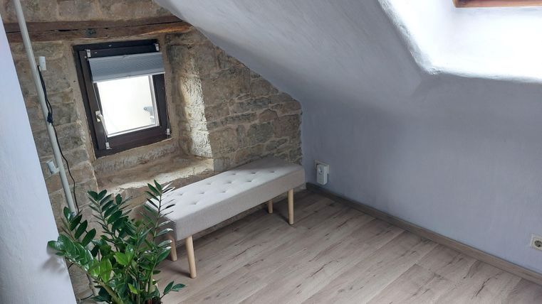 Een lichte kamer met een stenen muur en schuine wanden. Een smalle, lichte bank staat naast een raam.