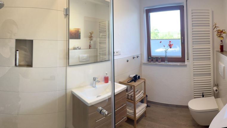 Een moderne badkamer met een douche en een groot raam. De muren zijn licht, en er zijn decoratieve elementen zoals bloemen en foto's.