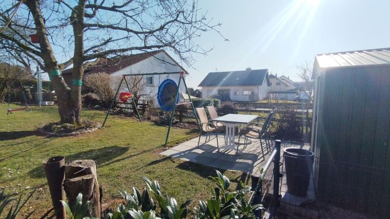 Ein ruhiger Garten mit einer Schaukel und einer gemütlichen Sitzecke. Im Hintergrund sind mehrere Häuser und ein klarer blauer Himmel zu sehen.