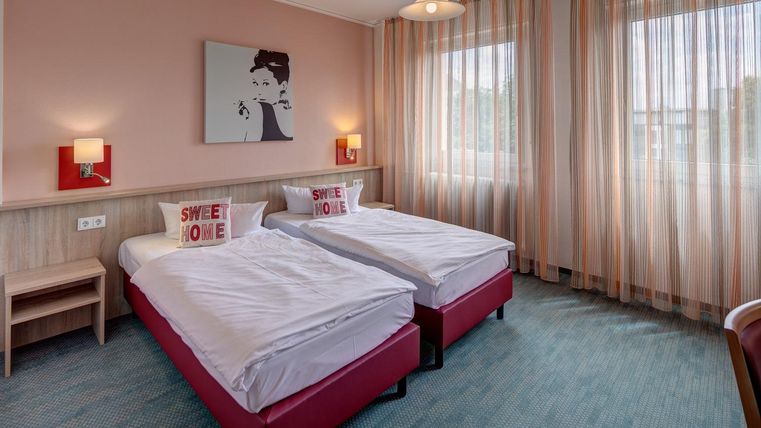 Ein modernes Schlafzimmer mit zwei Einzelbetten und einer warmen Farbgestaltung. Große Fenster mit Vorhängen sorgen für viel Tageslicht.