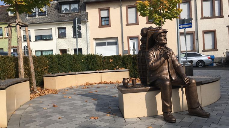 Une sculpture en bronze d'un homme est assise sur un banc. Autour de lui, on peut voir des arbres et des bâtiments modernes.