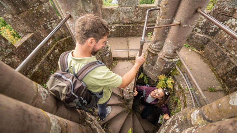 Deux personnes montent un escalier en colimaçon raide et en pierre. Une personne aide l'autre dans sa montée dans une vieille tour.