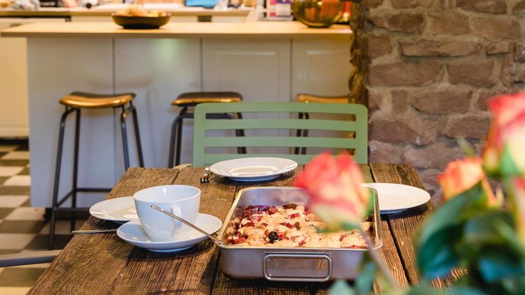 Ein einladender Esstisch mit einem Auflauf und gedeckten Tellern. Im Hintergrund ist eine moderne Küche mit Holzregalen zu sehen.