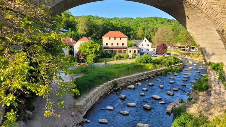 Ein malerischer Fluss fließt unter einer Steinbrücke. Im Hintergrund sind einige gemütliche Häuser und grünes Uferland zu sehen.