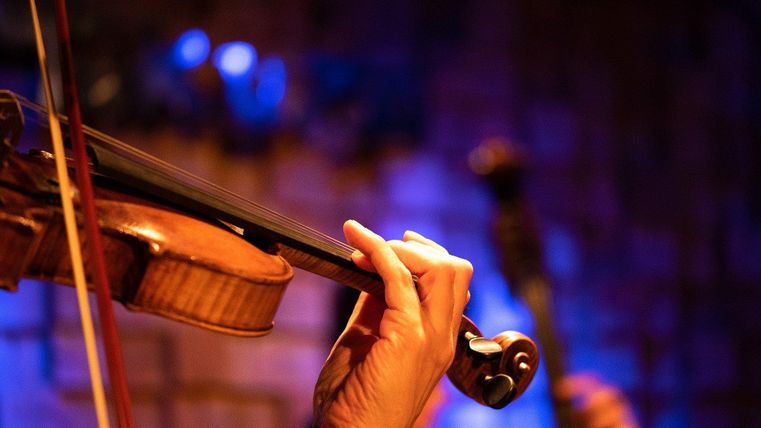 A hand is playing a violin on a stage. In the background, blurred lights are visible.
