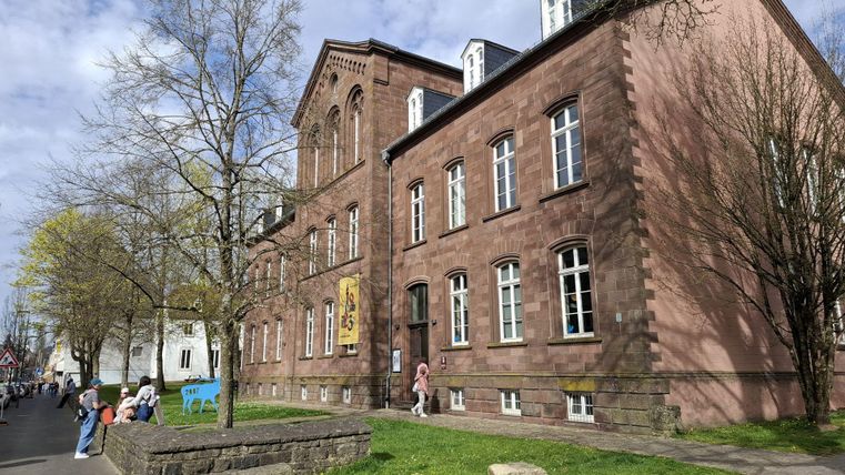 Een historisch gebouwencomplex van rode zandsteen met grote ramen. Op de voorgrond zijn bomen en enkele voorbijgangers te zien.