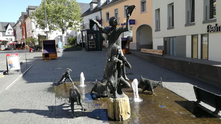 Eine moderne Statue steht auf einem gepflasterten Platz. Rundherum sprudeln kleine Brunnen, und einige Hunde sind ebenfalls abgebildet.