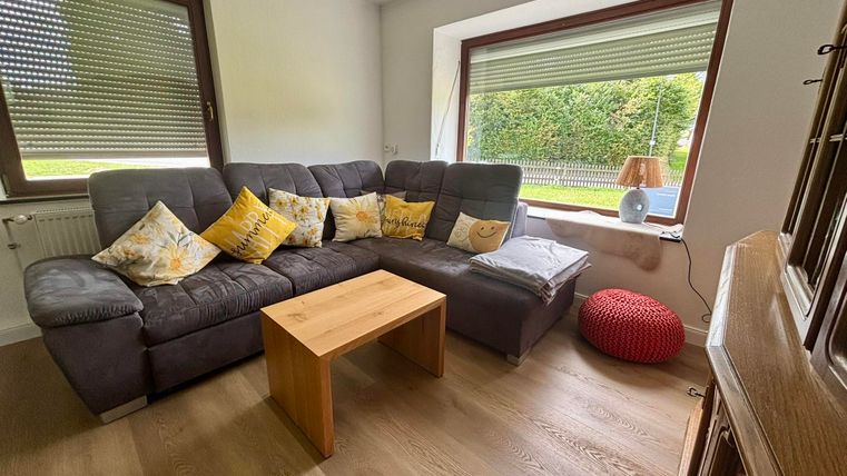 Un salon confortable avec un canapé gris et des coussins colorés. Une table en bois se trouve au centre et une fenêtre offre une vue sur le jardin.