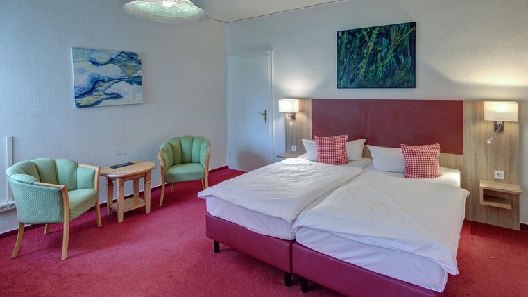 Ein gemütliches Hotelzimmer mit einem Doppelbett und einem kleinen Tisch. Die Wände sind hell, und es gibt zwei bequeme Sessel.