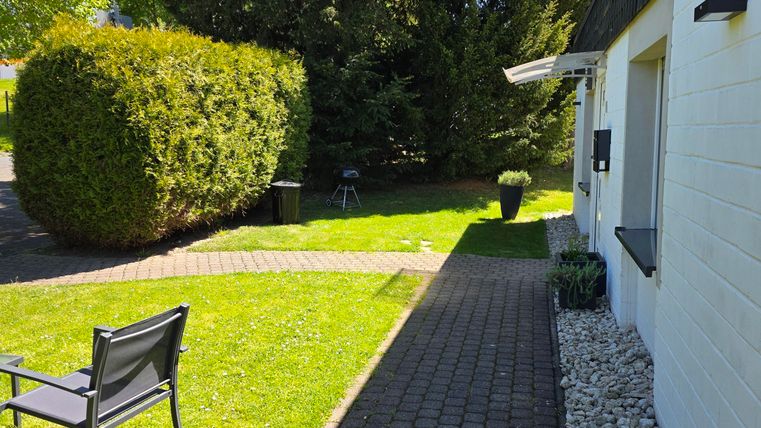 A well-maintained garden with green grass and bushes. On the path stands a garden chair and the sun is shining.
