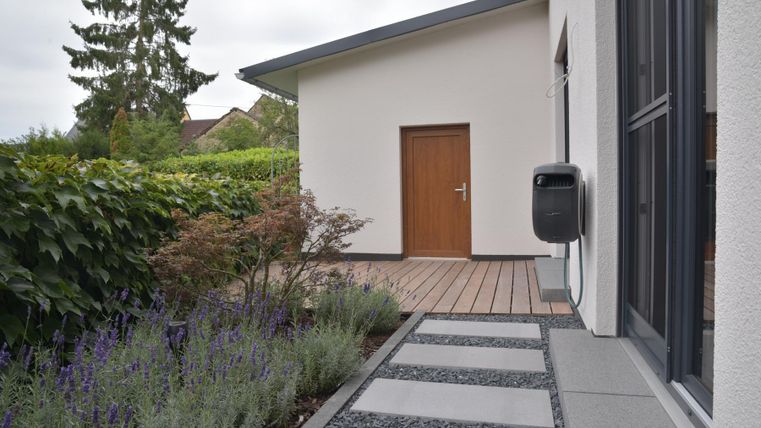 Ein moderner Eingangsbereich mit einem klaren Gehweg aus Steinen und Pflanzen. Im Hintergrund befindet sich ein Haus mit einer braunen Tür und grüner Bepflanzung.