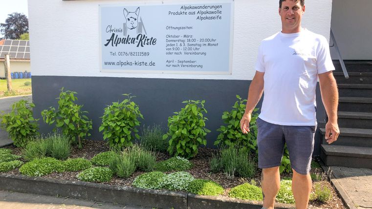 Ein Mann steht vor einem Gebäude mit einer weißen Fassade. Er trägt ein T-Shirt und Shorts, und um ihn herum sind grüne Pflanzen angeordnet.