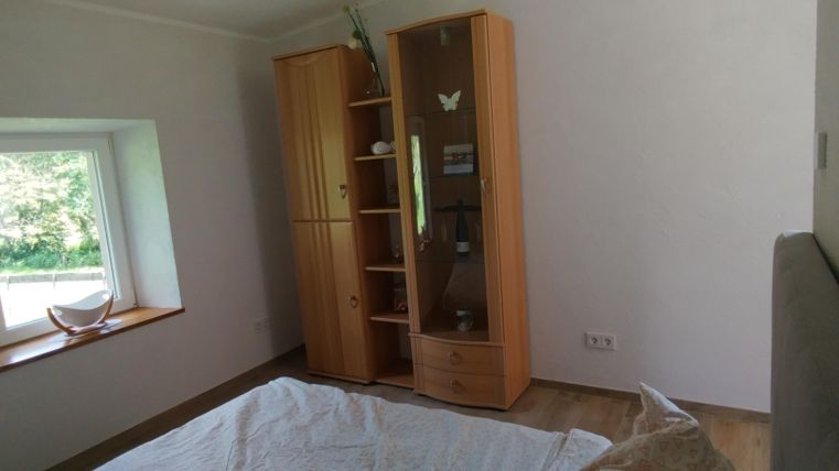 Ein einfaches Zimmer mit einem Holzschrank und einem Fenster. Der Boden ist aus Holz und das Bett hat eine weiße Bettdecke.