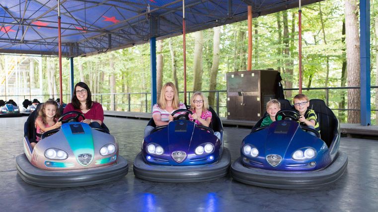 Eine Gruppe von Kindern und Erwachsenen sitzt in Autoscootern. Die Fahrzeuge stehen auf einer glatten Fläche unter einem überdachten Bereich.