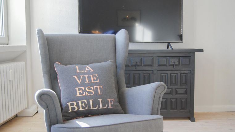 Ein bequemer Sessel mit einem Kissen, das „LA VIE EST BELLE“ sagt. Im Hintergrund steht ein Fernseher und ein dunkles Sideboard.