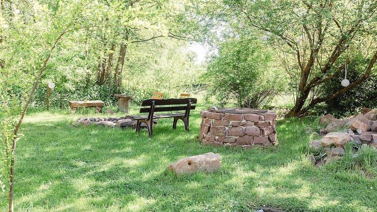 Ein ruhiger Garten mit grüner Wiese und schattenspendenden Bäumen. Eine Holzbank und ein gemauerter Feuerplatz laden zum Verweilen ein.