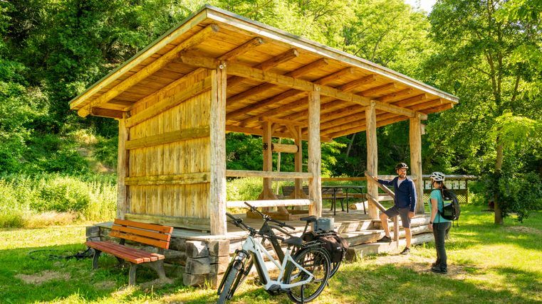Zwei Radfahrer stehen neben einem hölzernen Unterstand im Grünen.