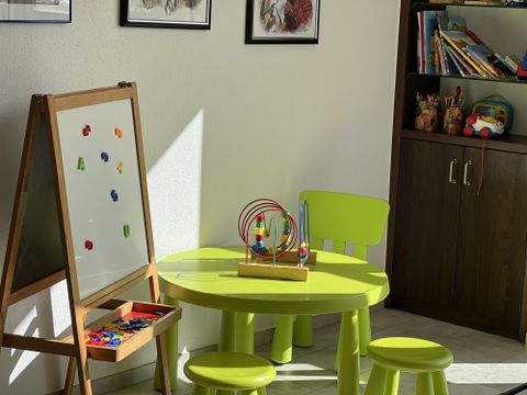 Ein farbenfrohes Kinderzimmer mit einem grünen Tisch und Stühlen. An der Wand steht eine Maltafel und ein Regal mit Spielsachen.