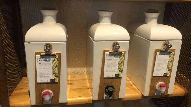 Drei weiße Behälter auf einem Regal, die mit Öl oder Essig gefüllt sind. Jeder Behälter hat ein Etikett und einen Auslauf.