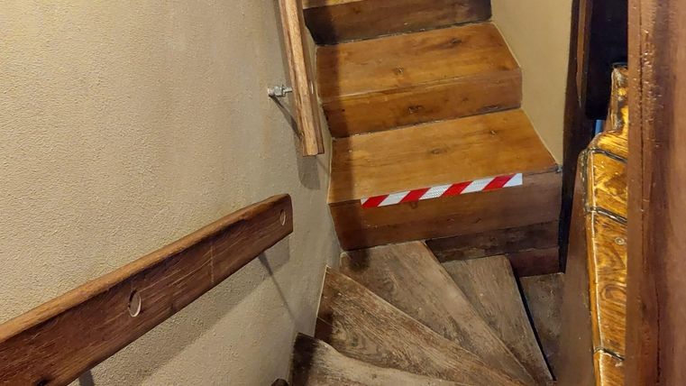 Een smalle, gedraaide houten trap leidt naar beneden. Aan de trap bevindt zich een veiligheidsmarkering.