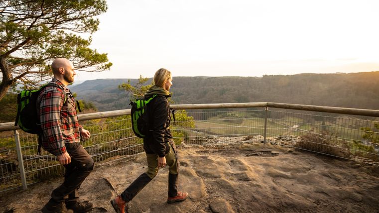 Zwei Wanderer mit Rucksäcken gehen auf einem Felsen, der einen schönen Ausblick bietet. Im Hintergrund ist eine malerische Landschaft bei Sonnenuntergang zu sehen.