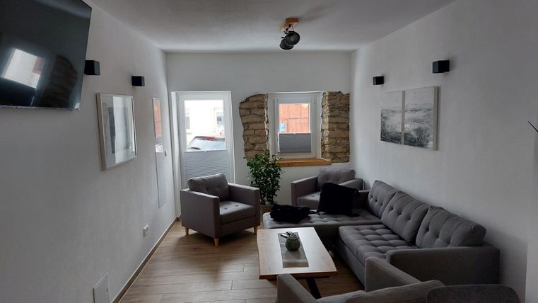 Een gezellige woonkamer met een grijze bank en stoelen. Aan de wand staan een planten en een televisie, het licht is helder en uitnodigend.