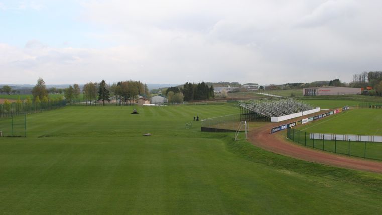 Eine große grüne Sportfläche mit Rasen und einem angrenzenden Stadion. Im Hintergrund sind einige Bäume und Gebäude zu erkennen.