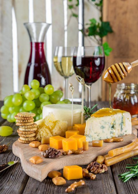 A stylish tray with various cheese types, honey, and nuts. In the background, wine glasses and grapes are visible.