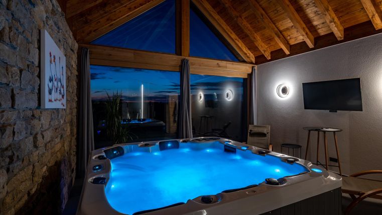 Ein moderner Whirlpool unter einem offenen Dach mit Holzverkleidung. Sanftes blaues Licht und Aussicht auf den Abendhimmel sorgen für eine entspannte Atmosphäre.