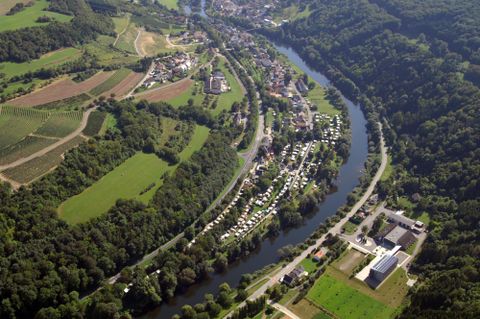 Eine Luftaufnahme eines malerischen Flusstals mit Häusern und Wiesen. Umgeben von grünen Hügeln und Wäldern fließt der Fluss ruhig durch die Landschaft.