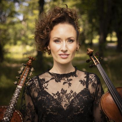 Eine Frau mit lockigem Haar steht im Freien und trägt ein schwarzes Kleid. Sie hält eine Geige und ein Cello in der Hand.