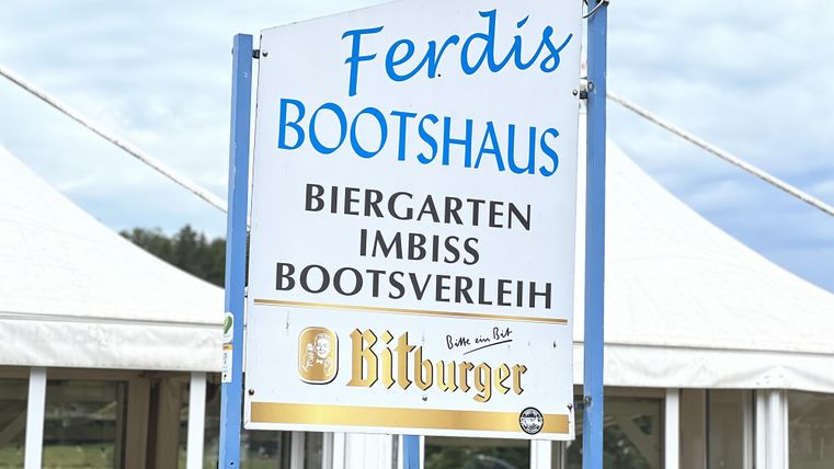 Ein Schild für das Ferdis Bootshaus, das einen Biergarten, einen Imbiss und Bootsverleih anbietet. Die Umgebung wirkt einladend und freundlich.