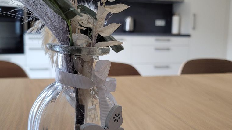 Eine Glasvase mit Trockenblumen und einer weißen Schleife steht auf einem Holztisch. Im Hintergrund ist eine moderne Küche zu sehen.