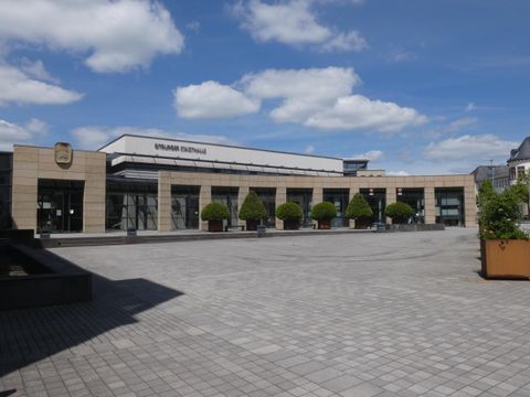 Un bâtiment moderne avec de grandes fenêtres et un ciel clair. La place devant le bâtiment est aménagée avec de jolis arbres et des jardinières.