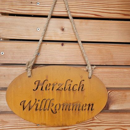 Ein hölzernes Willkommensschild hängt an einer Wand aus Holzbrettern. Der Text "Herzlich Willkommen" ist schön eingraviert.