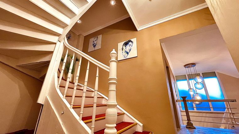 An elegant staircase with a white railing and red carpets leads upwards. Pictures are hung on the walls, and large windows can be seen in the background.