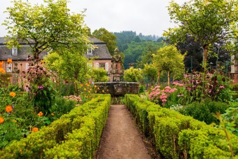 Ein schöner Garten mit bunten Blumen und gepflegten Hecken. Im Hintergrund sind Bäume und ein charmantes Gebäude sichtbar.
